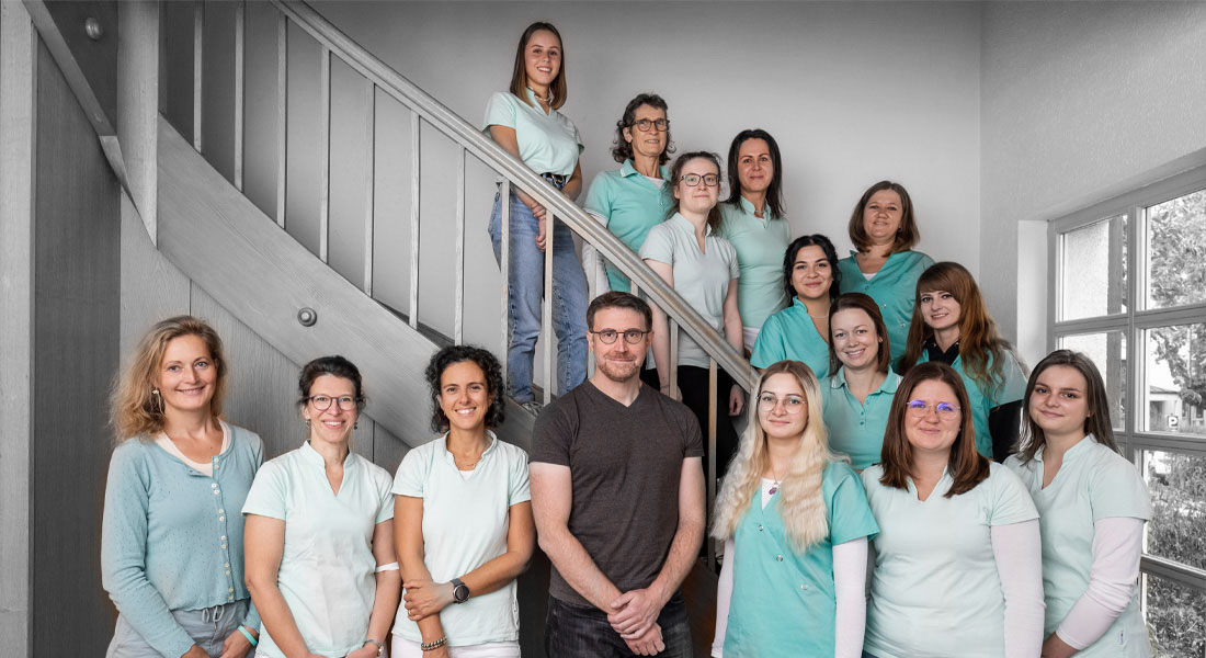 frauenaerzte-loerrach-schopfheim-team Das ganze Parxsteam, posieren lächelnd für ein Gruppenfoto in einem Treppenhaus.