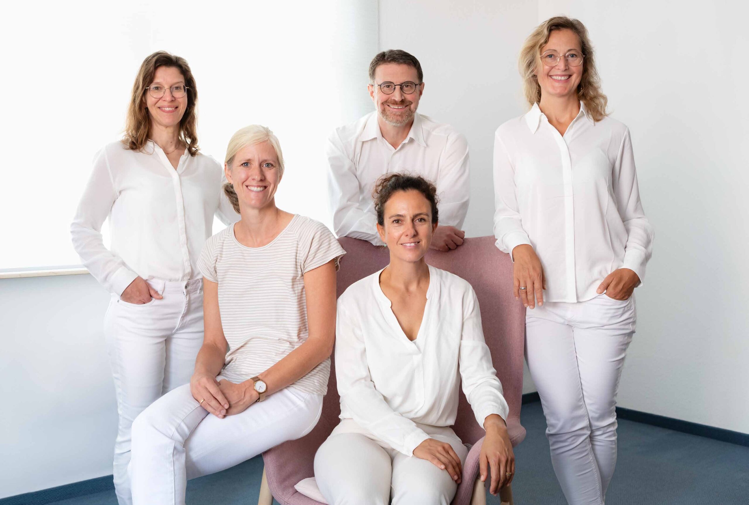 LY10_Website_Frauenarztpraxis-Loe-Sch Fünf Personen, alle in Weiß gekleidet, posieren lächelnd für ein Gruppenfoto in einem hellen Raum.